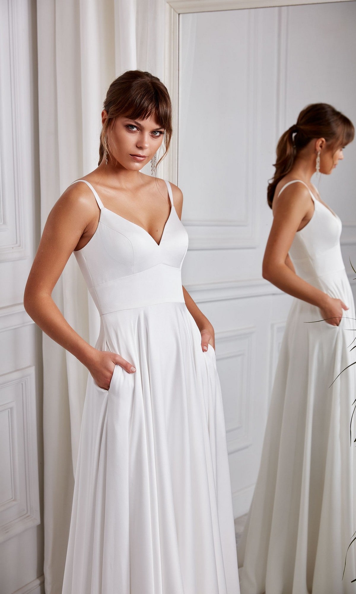A woman wearing the Aerie Couture 2003 Long White Dress with pockets and a v-neckline stands indoors by a mirror, looking at the camera.